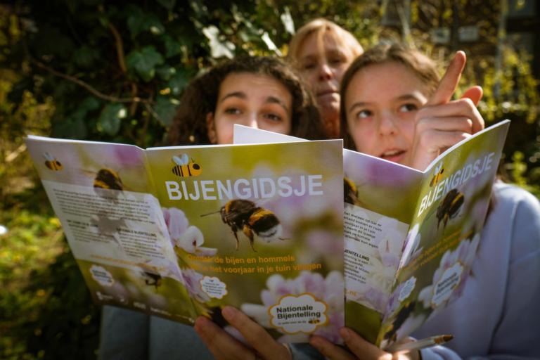 2 meisjes en een dame staan buiten achter het bijengidsje. het rechter meisje wijst naar iets buiten het scherm, de andere twee personen kijken naar de gids